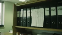 A Canadian office bookshelf with a row of binders labelled by year 2023 2024 2025 2026 and a printed hardware-amortization schedule paper-clipped to one binder