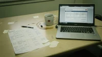 A Canadian conference table with a laptop angled away showing a non-readable cost calculator beside a printed inputs worksheet and a coffee mug